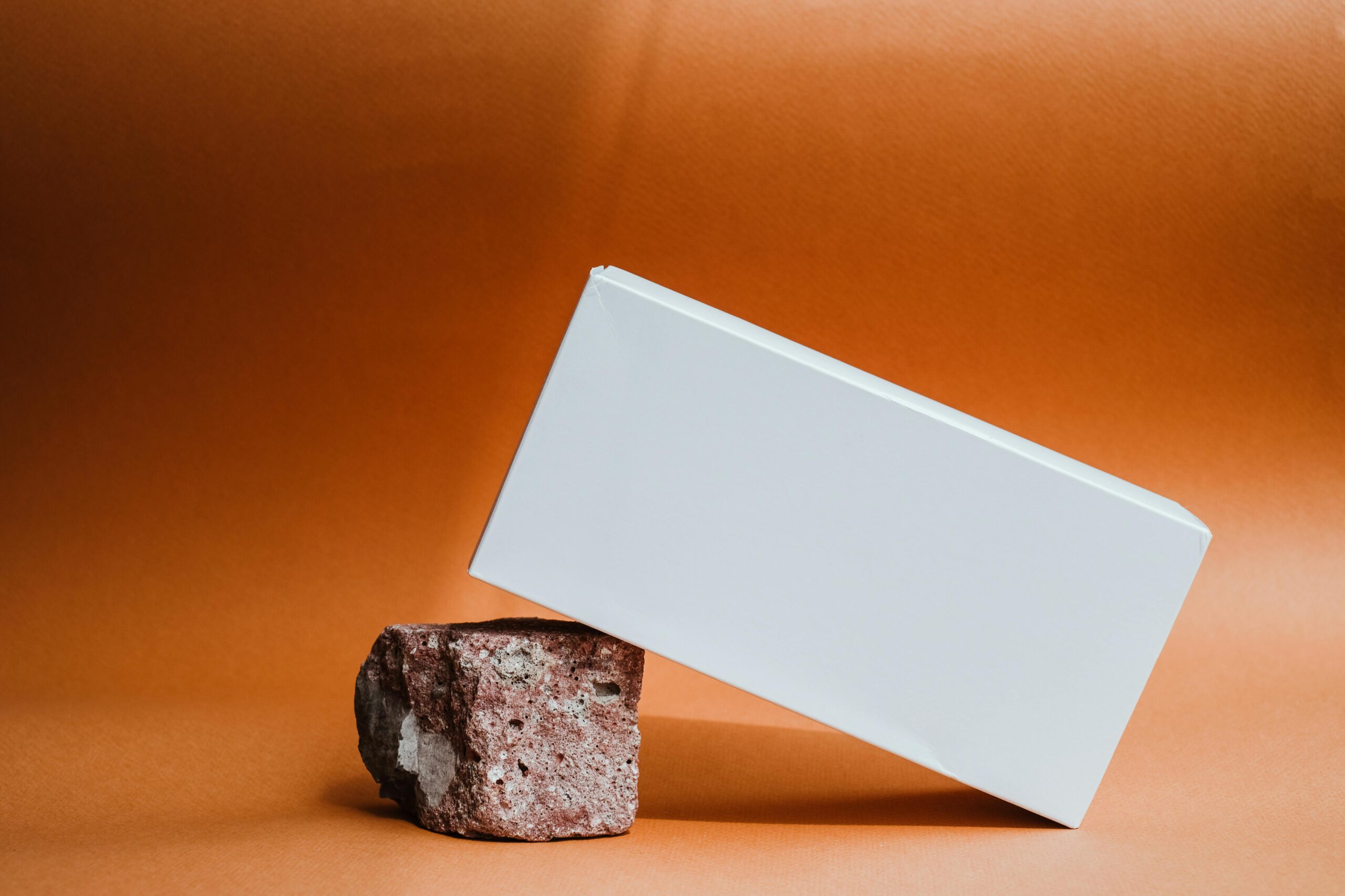 A minimalist still life of a white box perched on a brick against an orange backdrop.